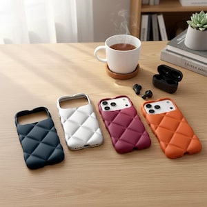 May include: Four quilted phone cases in navy, white, burgundy, and orange. The cases are arranged on a wooden surface with a cup of tea, a coaster, wireless earbuds, and a charging case.