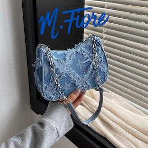 Puede incluir: Bolso de mano de mezclilla azul claro con un estampado de estrellas y una correa de cadena plateada. El bolso tiene un diseño deshilachado y un asa corta. El texto "M. Fiore" está escrito en azul en la parte superior izquierda.