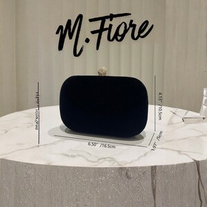 May include: A black velvet clutch with a rounded rectangular shape. The clutch has a jeweled clasp and measures 6.50 inches (16.5cm) wide, 4.13 inches (10.5cm) high, and 1.97 inches (5cm) deep. The name "M. Fiore" is written above the clutch.