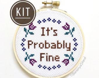 May include: A circular wooden embroidery hoop containing white Aida fabric with a cross-stitch design. The text reads It's Probably Fine in brown thread, surrounded by a circular pattern of small blue crosses and four purple and teal flowers.