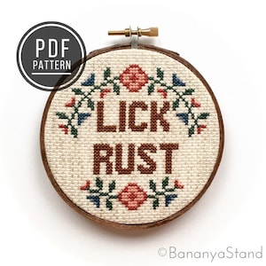 May include: A cross-stitch embroidery hoop with a beige fabric background and the words "LICK RUST" stitched in brown thread. The hoop is decorated with a floral pattern in shades of red, green, and blue.