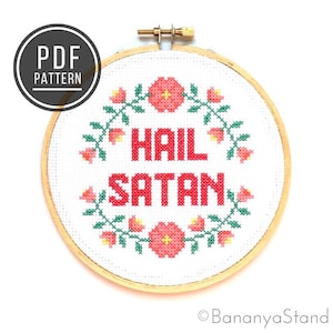 May include: A white embroidery hoop with a cross-stitch design featuring the phrase "Hail Satan" in red lettering surrounded by a wreath of pink flowers and green leaves.