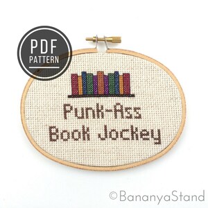 May include: A cross-stitch pattern of a bookshelf with colourful books and the text "Punk-Ass Book Jockey".