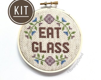 Puede incluir: Un kit de bordado de punto de cruz con el texto EAT GLASS en hilo marrón sobre un fondo de tela beige. El diseño está enmarcado en un bastidor de madera circular y decorado con pequeños motivos florales morados y azules sobre y bajo el texto.