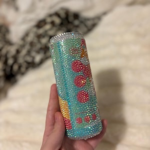 May include: A bedazzled tumbler with a turquoise and pink design. The tumbler is covered in iridescent rhinestones, with a pattern of pink circles and yellow accents. The rim of the tumbler is also lined with rhinestones.