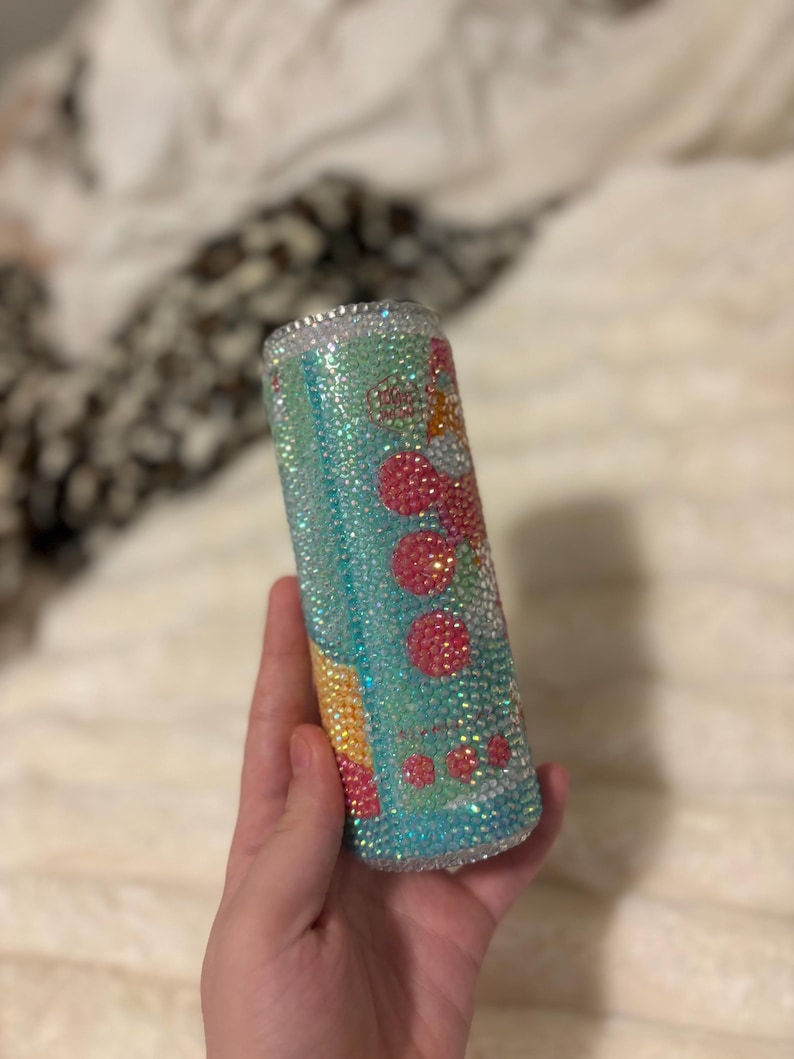 May include: A bedazzled tumbler with a turquoise and pink design. The tumbler is covered in iridescent rhinestones, with a pattern of pink circles and yellow accents. The rim of the tumbler is also lined with rhinestones.