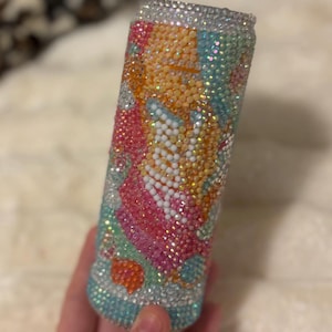 May include: A bedazzled tumbler with a colorful design. The tumbler is covered in small, sparkling rhinestones in shades of blue, pink, orange, and white. The design appears to be floral or abstract. The top and bottom rims are also covered in rhinestones.