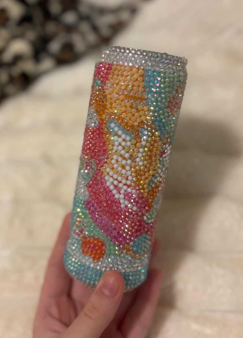 May include: A bedazzled tumbler with a colorful design. The tumbler is covered in small, sparkling rhinestones in shades of blue, pink, orange, and white. The design appears to be floral or abstract. The top and bottom rims are also covered in rhinestones.