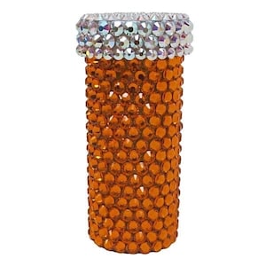 May include: An orange, bedazzled pill bottle with a silver, crystal-studded top. The bottle is covered in small, orange crystals, creating a sparkling effect. The design is eye-catching and decorative.