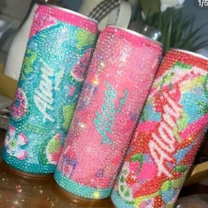 May include: Three bedazzled beverage cans in shades of pink, blue, and green. Each can is covered in sparkling rhinestones, with the word "Alan" in cursive. The cans have pink rims and are decorated with colorful patterns.