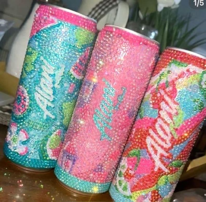 May include: Three bedazzled beverage cans in shades of pink, blue, and green. Each can is covered in sparkling rhinestones, with the word "Alan" in cursive. The cans have pink rims and are decorated with colorful patterns.