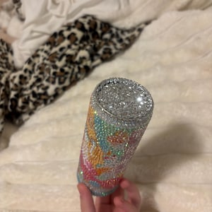 May include: A bedazzled tumbler with a silver rim and a colorful design. The tumbler is covered in small, sparkling rhinestones in shades of blue, pink, yellow, and green. The design is abstract and swirly, with a silver rhinestone top.