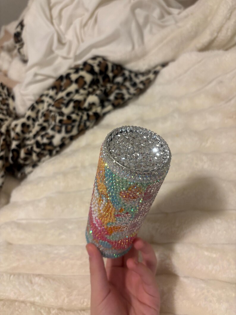 May include: A bedazzled tumbler with a silver rim and a colorful design. The tumbler is covered in small, sparkling rhinestones in shades of blue, pink, yellow, and green. The design is abstract and swirly, with a silver rhinestone top.