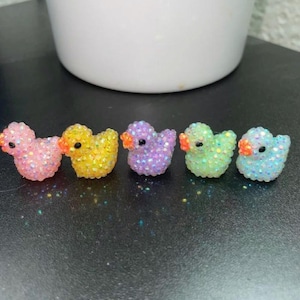 May include: A row of five glittery duck figurines in pink, yellow, purple, green, and blue. Each duck has an orange beak and black eyes. The ducks are arranged on a dark surface, with a white pot in the background.