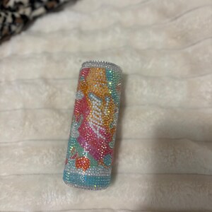 May include: Cylindrical container covered in colorful rhinestones. The rhinestones are arranged in a pattern of orange, pink, white, and blue. The top and bottom edges are lined with silver rhinestones. The container is resting on a white, textured surface.