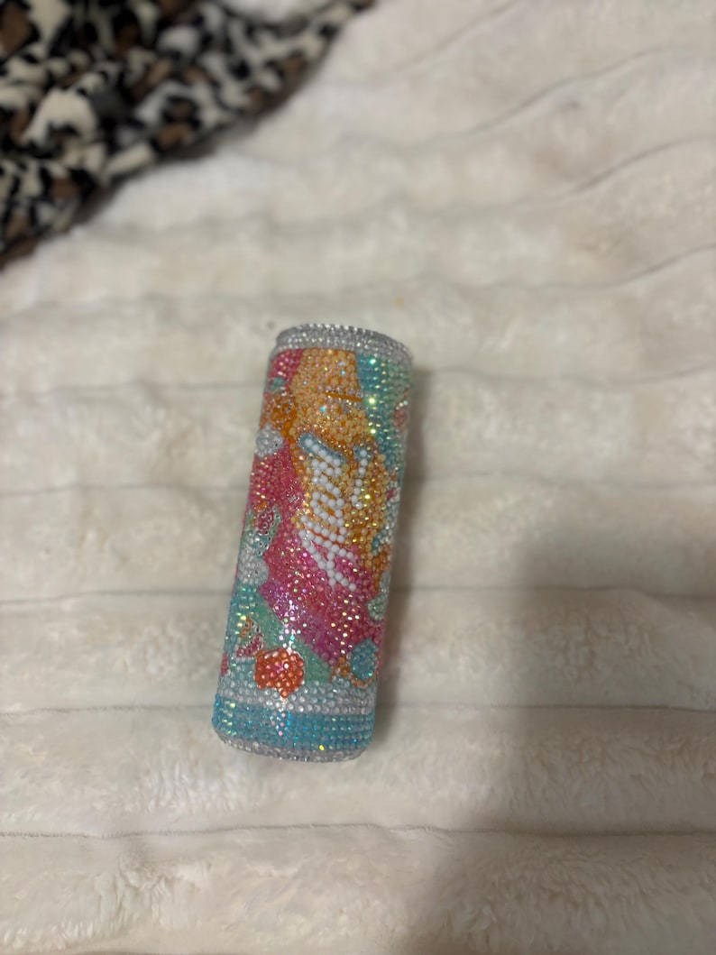 May include: Cylindrical container covered in colorful rhinestones. The rhinestones are arranged in a pattern of orange, pink, white, and blue. The top and bottom edges are lined with silver rhinestones. The container is resting on a white, textured surface.