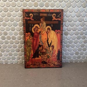 May include: A rectangular religious icon depicting a scene with figures around a cross. The artwork features a dark background with figures in red, yellow, and green robes. The icon has text at the top and is framed with decorative borders.