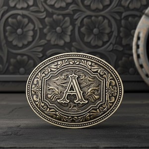 Puede incluir: Hebilla de cinturón ovalada de latón antiguo con la letra "A" en el centro. La hebilla presenta un diseño floral y de desplazamiento ornamentado alrededor de la letra y el borde exterior. Una herradura es visible en el fondo.