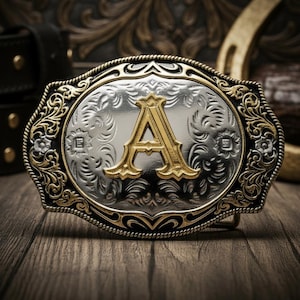 Op de afbeelding: Ovale zilver- en goudkleurige riemgesp met de gouden letter "A". De gesp heeft een sierlijk bloemmotief langs de randen en staat op een houten achtergrond. De gesp is waarschijnlijk van metaal.