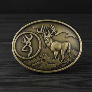 May include: Oval-shaped, antique brass belt buckle featuring a detailed relief of a deer with large antlers in a natural landscape. The buckle also includes a circular logo with a stylized deer head.