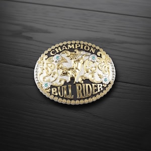 Pode incluir: Fivela de cinto ovalada prateada e dourada com as palavras "CHAMPION BULL RIDER". A fivela apresenta um design de cavaleiro de touro com detalhes turquesa e ornamentos ornamentados. A fivela tem uma borda dourada.