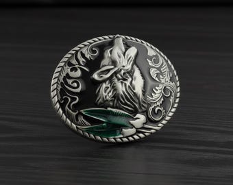 May include: Silver-toned metal belt buckle featuring a howling wolf design. The wolf is in the center, surrounded by ornate scrollwork and a green accent. The buckle is round and has a black background.