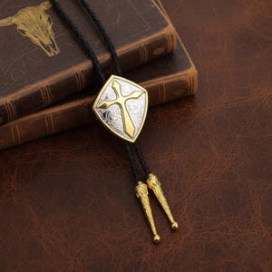 May include: A bolo tie with a silver and gold cross pendant. The pendant has a gold cross on a silver background with an ornate design. The black leather cord has gold-colored tips. The bolo tie is displayed on a leather surface.
