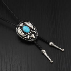 Op de afbeelding: Ovale bolo stropdas met een zilverkleurig metalen ontwerp en een turquoise gekleurde steen in het midden. De bolo stropdas heeft een zwart gevlochten leren koord met decoratieve zilveren uiteinden.