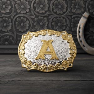 Puede incluir: Hebilla de cinturón de metal plateado y dorado con la letra "A" en oro. La hebilla tiene un diseño floral ornamentado en los bordes y en el fondo plateado. Una herradura es visible en el fondo.