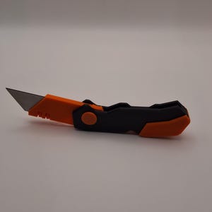 May include: A utility knife with a retractable blade. The handle is black and orange, with a circular button for blade control. The blade is silver and angled, designed for cutting. The knife is shown on a white background.