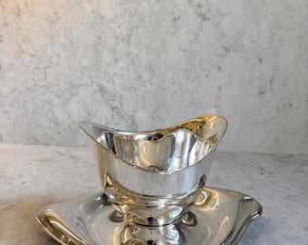 May include: A silver gravy boat and matching saucer set. The boat has a curved rim and a handle-like spout. The saucer is a similar shape, with a slightly raised edge. The set is highly reflective, with a polished finish.