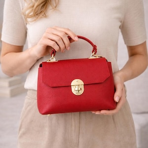 May include: A red leather handbag with a top handle and a gold-colored clasp. The bag has a rectangular shape and a textured surface. The handbag is held by a person wearing a cream-colored top and tan pants.