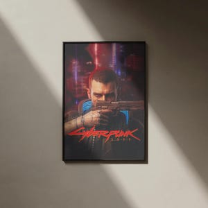 K&ouml;nnte beinhalten: Gerahmtes Poster mit einer Person, die eine Waffe h&auml;lt, und dem roten Text "Cyberpunk 2077". Der Hintergrund zeigt eine verschwommene Stadtlandschaft mit roten und violetten Farbt&ouml;nen. Das Poster ist in einem schwarzen Rahmen.