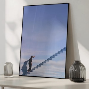 May include: Framed art print depicting a person climbing a staircase that extends into a blue sky. The artwork is in a black frame. Decorative vases are on either side of the framed print, displayed on a white surface.