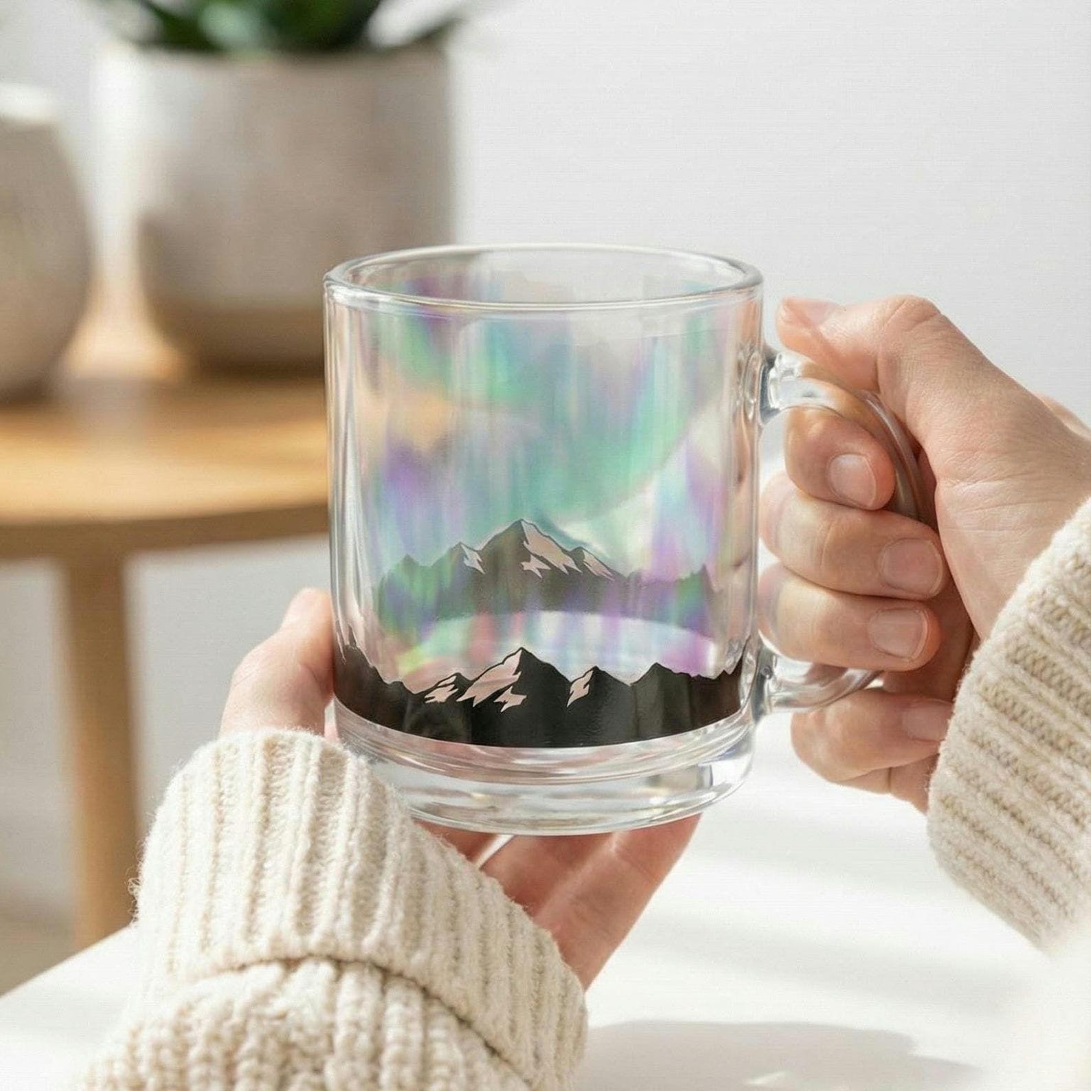 Two hands holding the aurora borealis glass mug up to the light, showing the iridescent mountain design