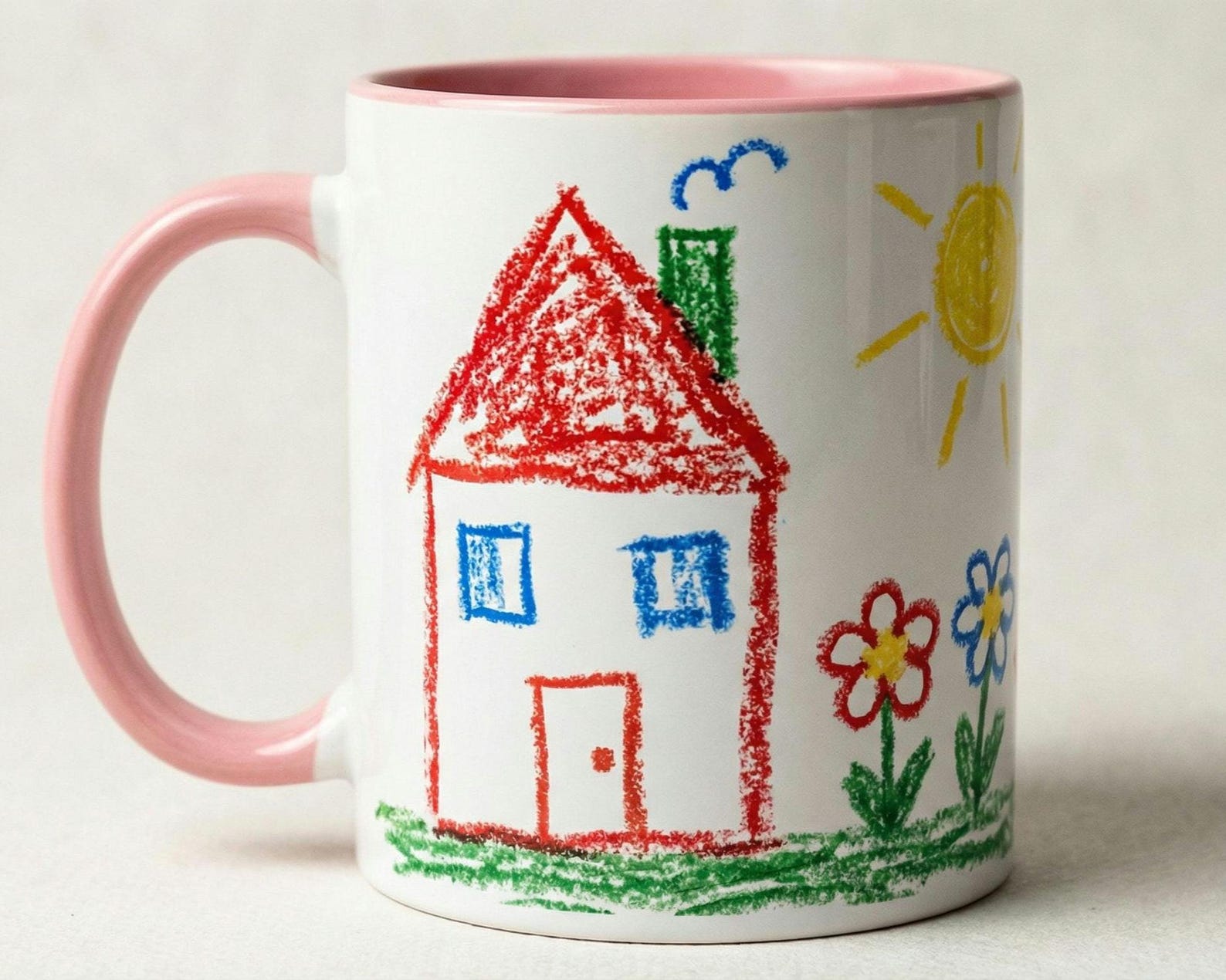 White mug with a pink interior and handle, printed with a child's crayon drawing of a house, sun, and flowers