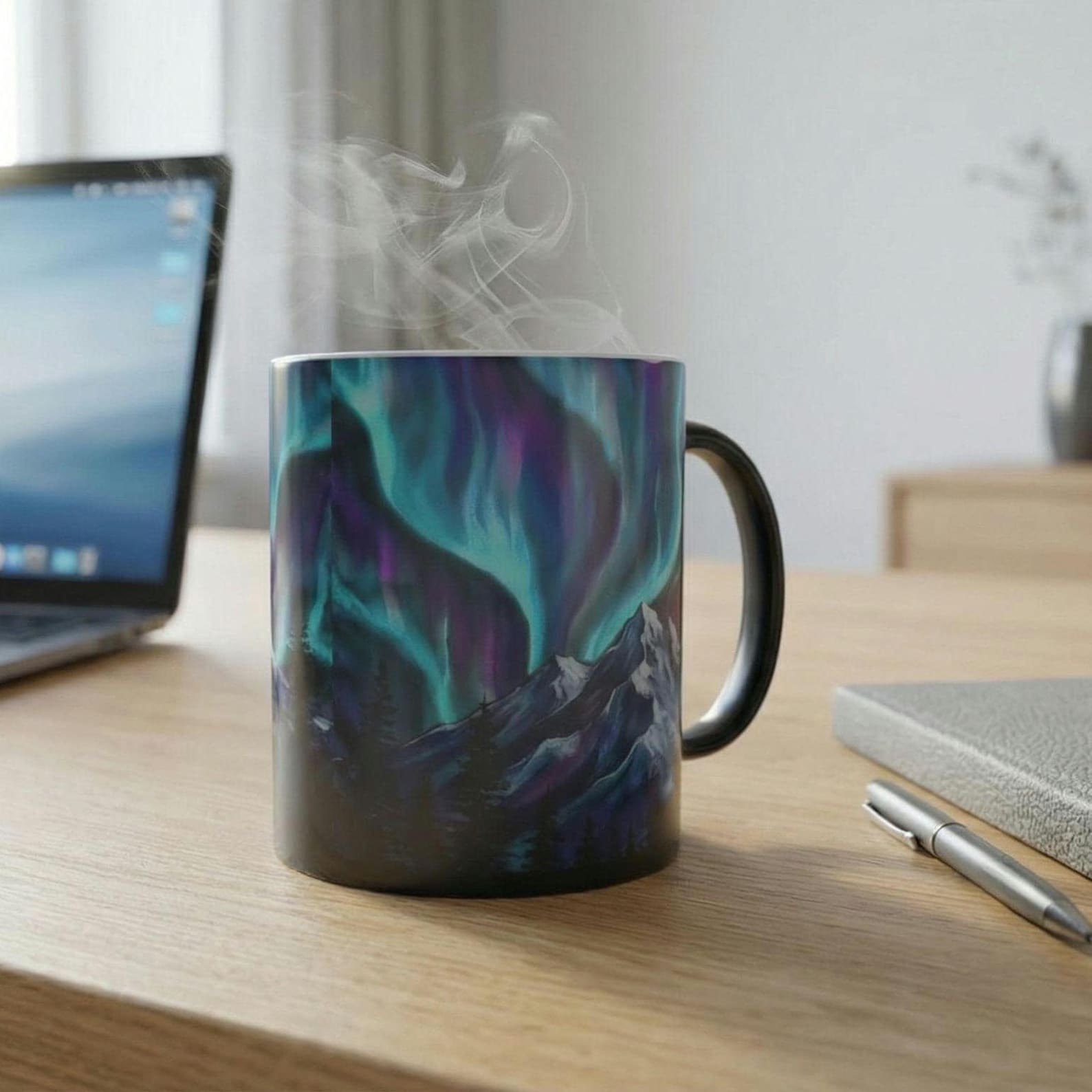 Close-up of the color-changing mug on a wooden desk beside a laptop and notebook, aurora borealis design fully revealed