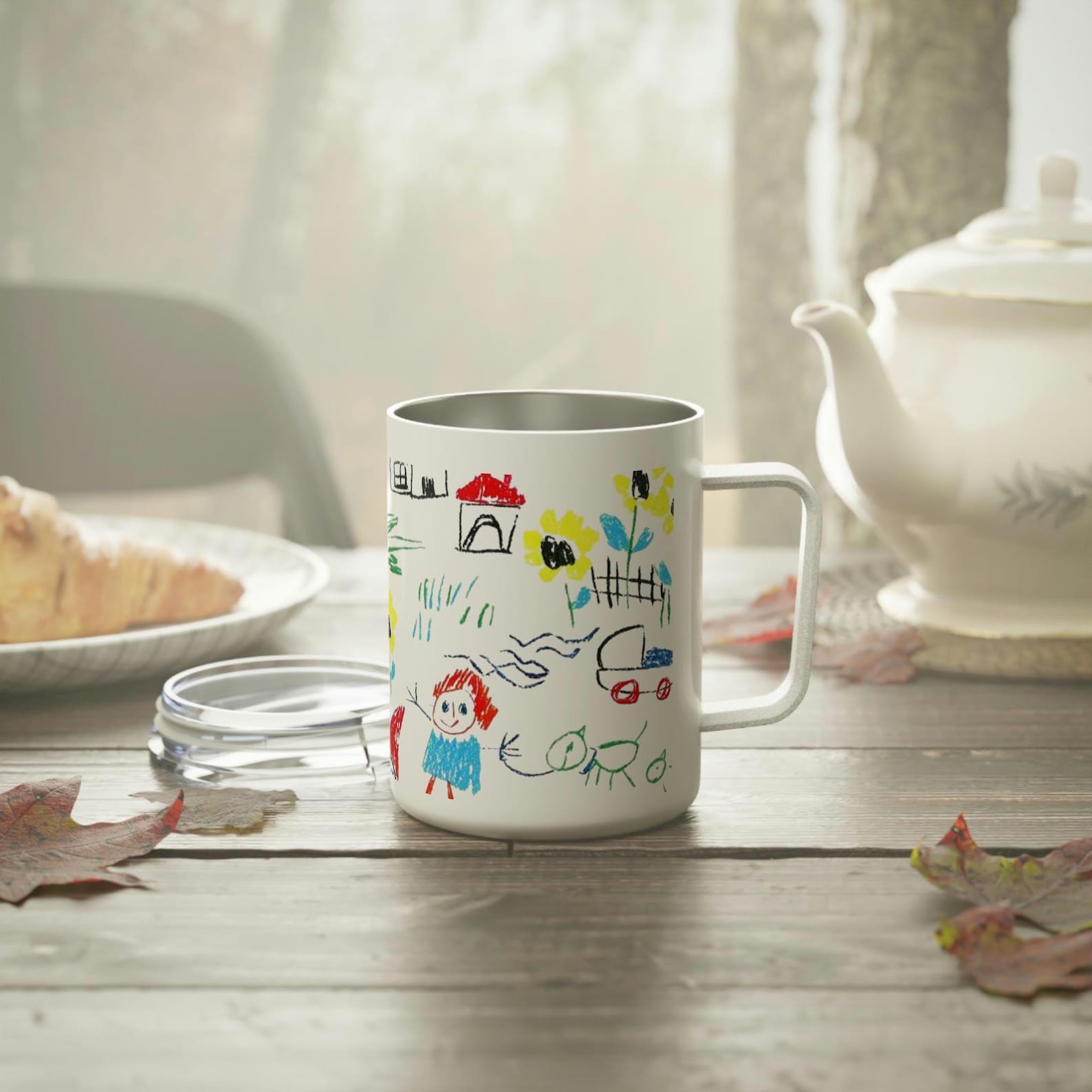 Insulated mug printed with a child's colourful drawing of a house, flowers, and family, on a table with a teapot and croissant