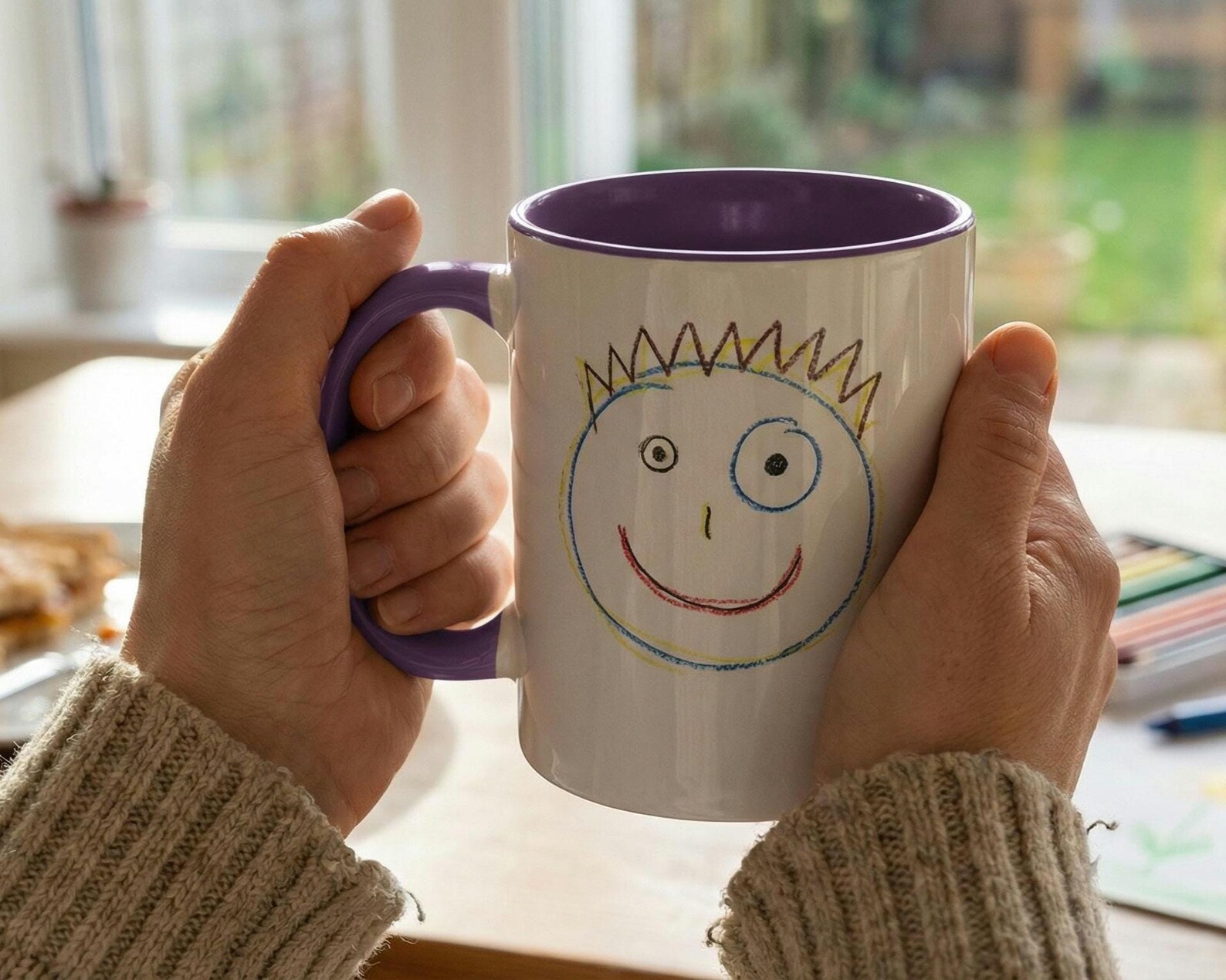 Two hands holding a white mug with a purple interior and handle, printed with a child's drawing of a smiling face