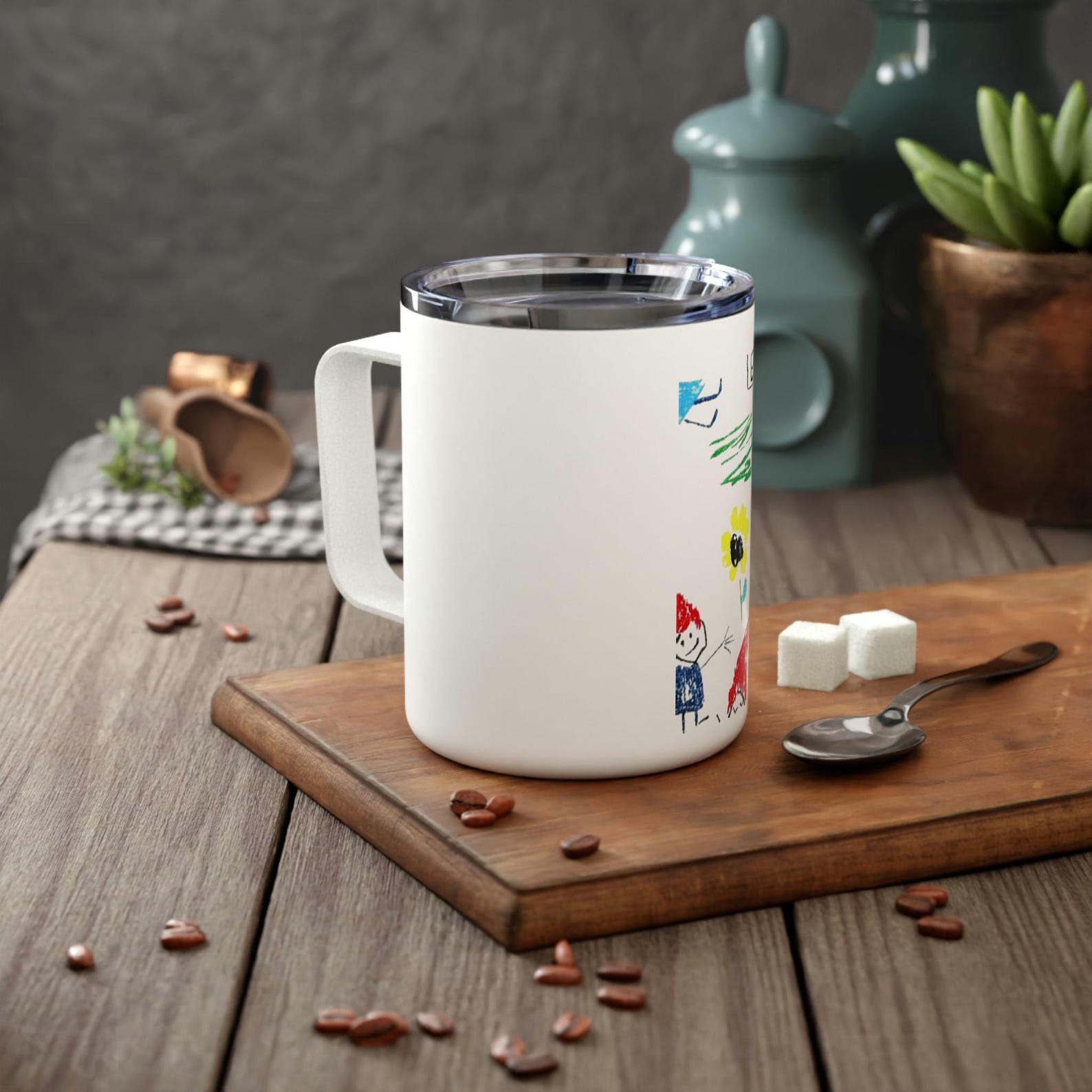 Insulated stainless steel mug with a clear lid, printed with a child's colourful drawing, on a wooden board with coffee beans and sugar cubes