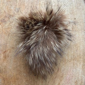 Può includere: Un soffice pompon in pelliccia marrone e beige. Il pompon ha un aspetto denso e strutturato con peli lunghi e sottili. I colori vanno dal marrone scuro al beige chiaro, creando un effetto naturale e stratificato. Il pompon è probabilmente un accessorio decorativo.