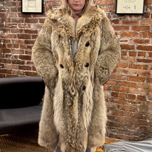 May include: A long, double-breasted fur coat in shades of brown and tan. The coat has large lapels and black buttons. The person is wearing blue jeans and white trainers. The background is a brick wall. The coat's texture is visible.