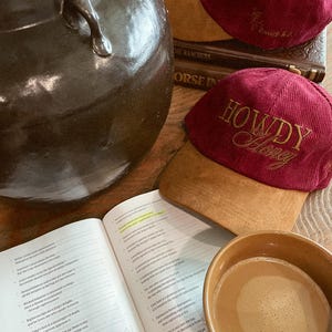 以下が含まれることがあります： 「HOWDY Honey」の金色の刺繍が入った、2つのバーガンディ色のコーデュロイベースボールキャップ、聖書、コーヒーカップ、大きな茶色のセラミックポットをフィーチャーした静物画。キャップにはタンのスエードバイザーが付いています。