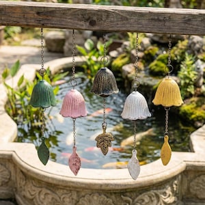 May include: A collection of six colorful bell-shaped wind chimes hanging from a wooden beam. The chimes are in shades of green, pink, bronze, white, and yellow. Each chime has a leaf-shaped clapper. The background features a pond with fish.