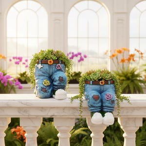 Puede incluir: Dos jardineras decorativas con forma de vaqueros de mezclilla, llenas de suculentas. Cada jardinera tiene un cinturón marrón, parches de rosas y pies blancos estilo zapatilla. Se exhiben sobre una repisa de mármol blanco, con un fondo borroso de flores.
