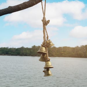 May include: A hanging wind chime with four brass bells suspended from a natural rope. A decorative butterfly and sun charm adorn the rope. The bells are hanging from a tree branch against a backdrop of a lake and sky.