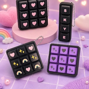 May include: Four black keychains with square and rectangular designs. One square keychain has pink hearts on the keys. Another has rainbows, stars, and hearts. The third has purple hearts and stars. The rectangular keychain spells "S K Y E".