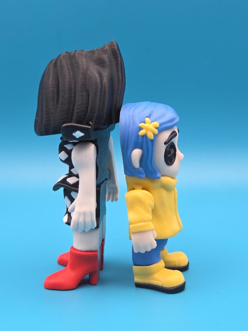May include: Two stylized figurines standing back-to-back. The left figure has black hair, a black dress with white diamond patterns, and red boots. The right figure has blue hair with a yellow flower accessory, a yellow jacket, and yellow boots.