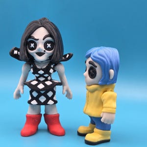May include: Two stylized figurines. The left figure has black hair, button eyes, and a black and white diamond-patterned dress with red boots. The right figure has blue hair, button eyes, a yellow hooded jacket, blue pants, and yellow boots.