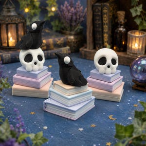 May include: Three stacks of miniature pastel-colored books serve as pedestals for two black raven figurines with white circular eyes and one white skull figurine with black eye sockets and a small nose cavity, arranged on a dark blue surface.